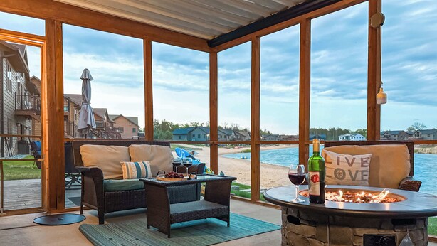 Une terrasse couverte, meublée de mobilier en osier, avec un foyer en pierre et une vue sur la plage et les maisons.