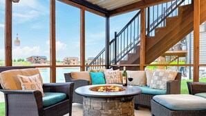Un espace extérieur aménagé, abrité et doté de mobilier en osier, d’une table avec foyer en pierre, et offrant une vue sur un lac et des bâtiments.