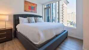 A modern bedroom with a large bed, a nightstand, a window with a city view, and a piece of abstract art on the wall. - Luxury Apt with Kitchen and Pool (Boston)