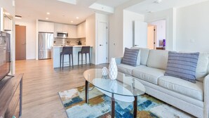 A modern living room with a glass coffee table, a sofa, and a kitchen in the background. - Luxury Apt with Kitchen and Pool (Boston)