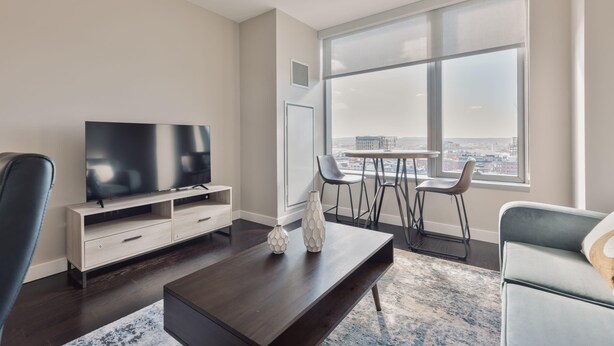 Ein modernes Wohnzimmer mit großem Fernseher, Couchtisch und Blick auf eine Stadtlandschaft durch große Fenster.