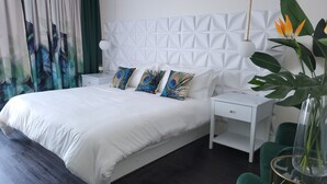 Ein modernes Schlafzimmer mit einem weißen Bett, Kissen mit Pfau-Motiv, einem weißen Nachttisch, einem grünen Sessel und einer großen Pflanze.