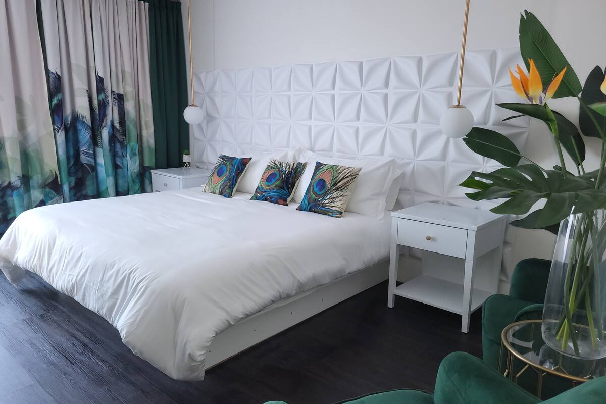 A modern bedroom with a white bed, peacock-themed pillows, a white nightstand, a green armchair, and a large plant.