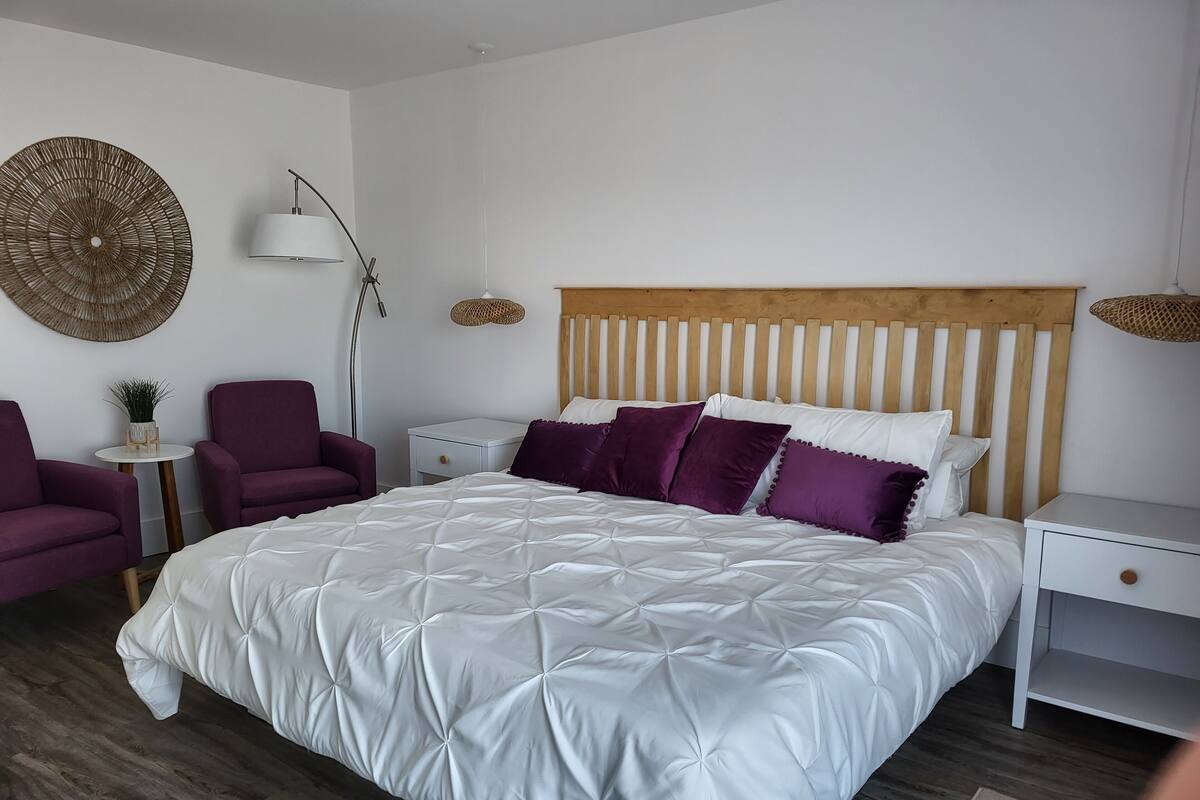 A bedroom with a large bed, two nightstands, a wooden headboard, a floor lamp, and two purple armchairs.