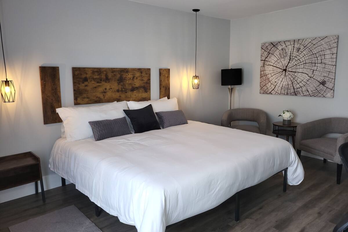 A modern bedroom with a large bed, wooden headboard, two wall-mounted lights, a side table with a vase, and a large abstract painting.