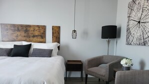 Ein modernes Schlafzimmer mit einem großen Bett, einem grauen Sessel, einem kleinen Beistelltisch, einer Stehlampe und einem abstrakten Kunstwerk an der Wand.