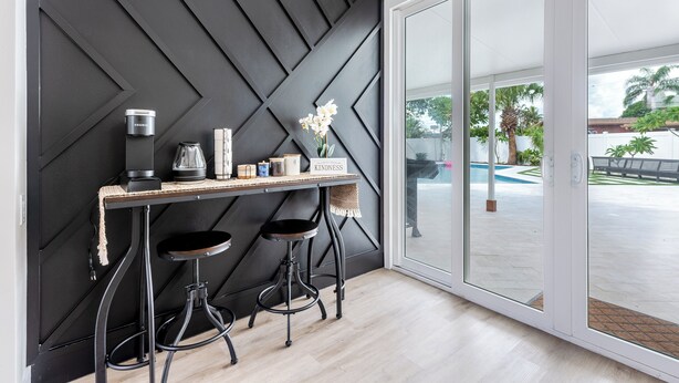Une cuisine moderne avec un mur noir à motifs géométriques, un plan de travail en bois, deux tabourets et une vue sur une piscine, visible à travers des portes vitrées.