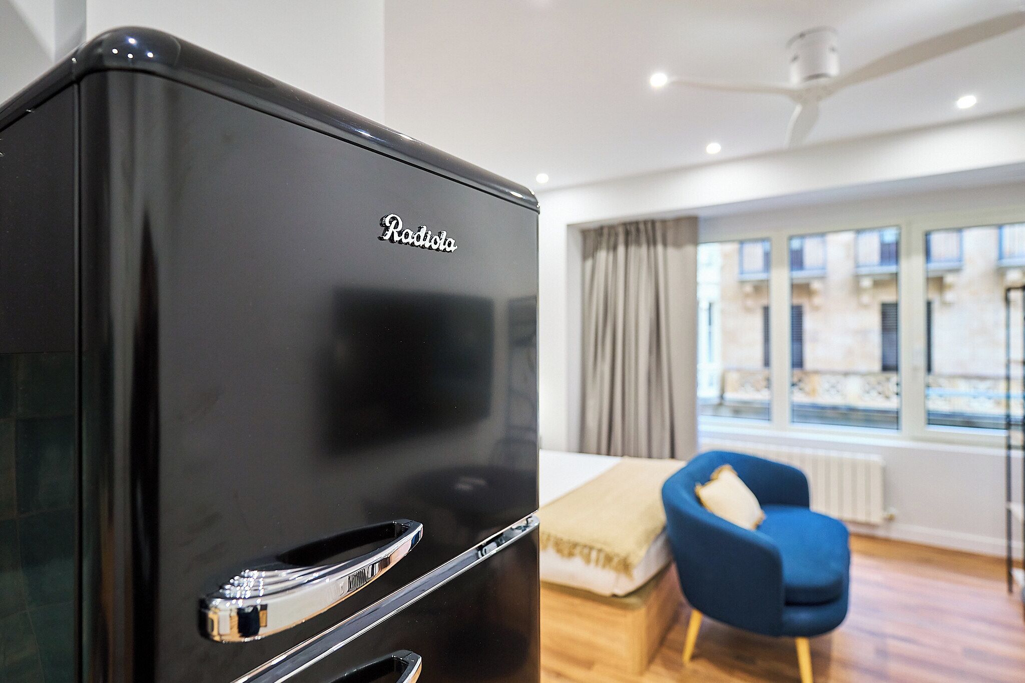 A black refrigerator with the brand name "Radiola" in a modern room with a blue chair, a bed, and a window with curtains.
