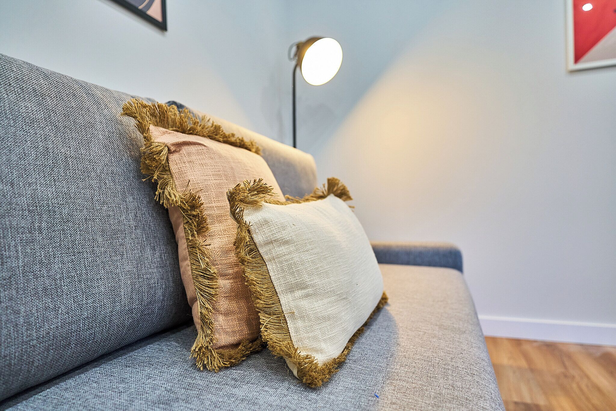 Un sofá gris con tres almohadones, uno beige y dos marrones, con detalles de flecos.