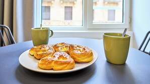 Een tafel met een bord gebakjes en twee gele bekers, een raam met uitzicht op gebouwen, en een gordijn.