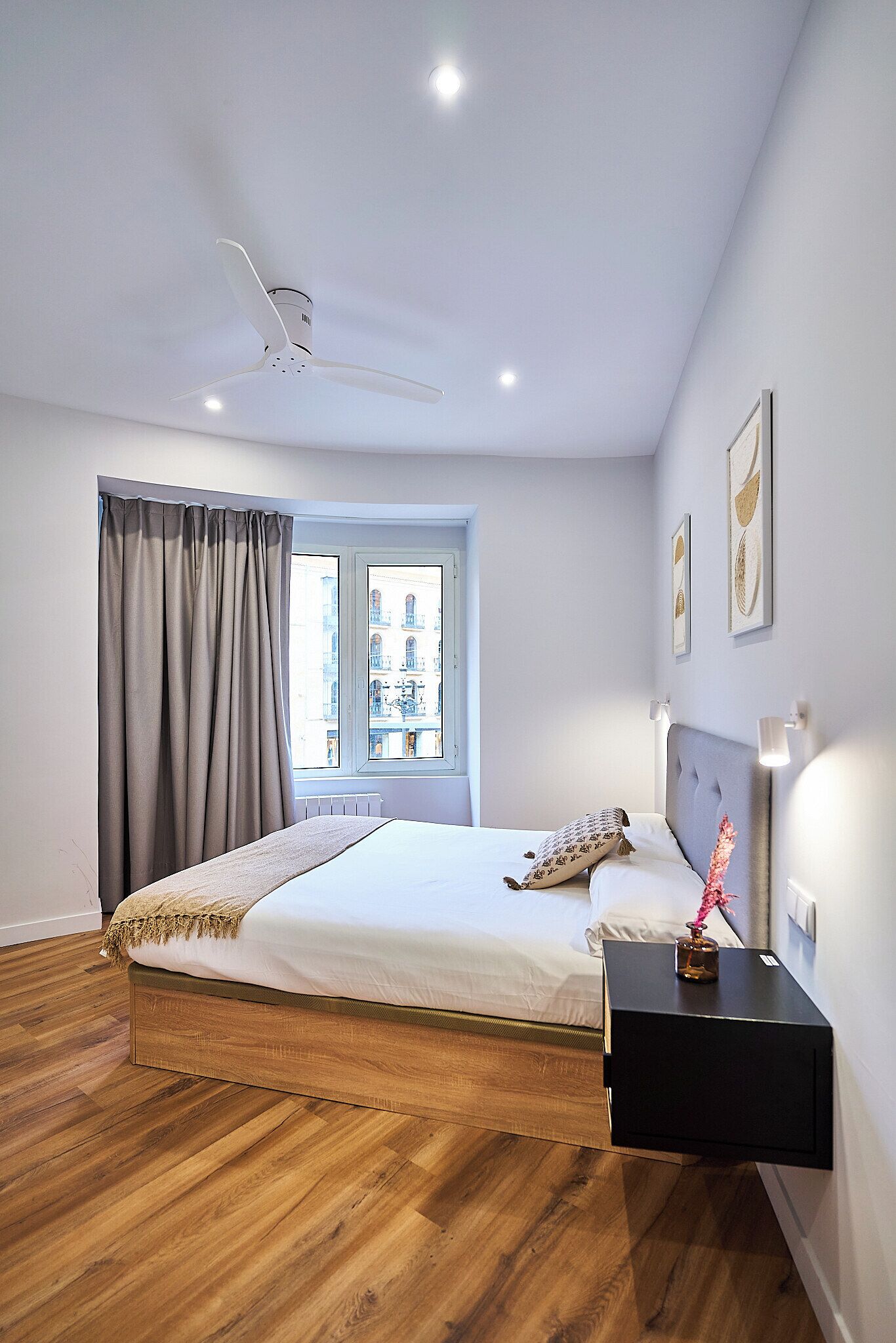 A modern bedroom with a wooden floor, a bed with a white mattress and a grey headboard, a bedside table with a lamp, and a window with curtains.