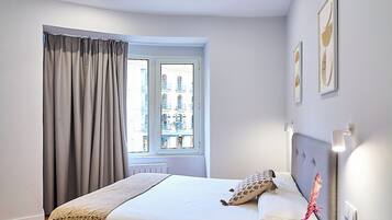 A modern bedroom with a wooden floor, a bed with a white mattress and a grey headboard, a bedside table with a lamp, and a window with curtains.