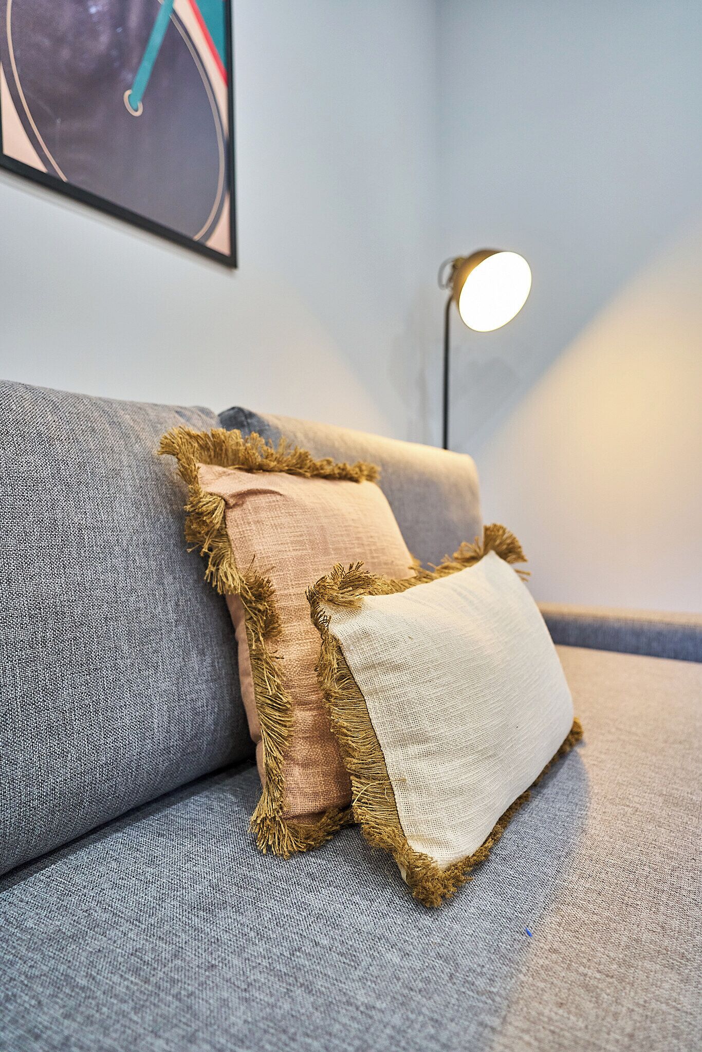 Un canapé gris avec deux coussins, dont l’un est orné de glands et l’autre est uni, dans une pièce où est fixée une lampe au mur.