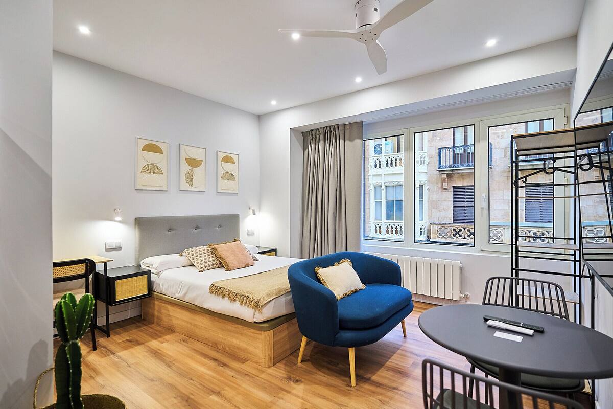 Une chambre à coucher moderne avec un lit, un fauteuil bleu, une petite table et un ventilateur de plafond.