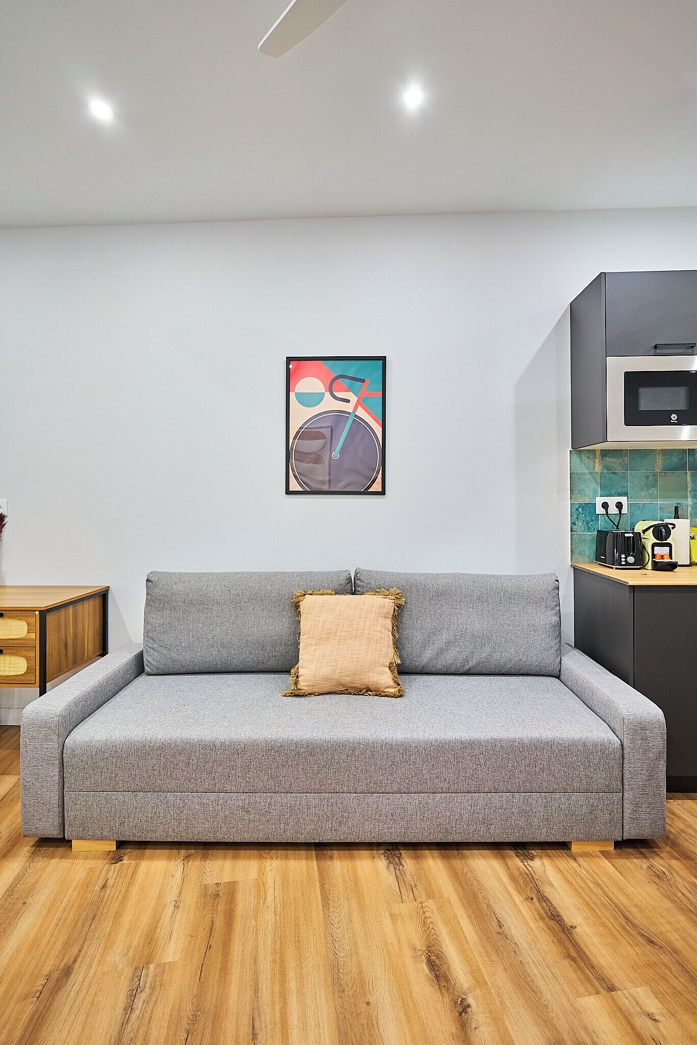 A modern living room with a grey sofa, a wooden floor, a kitchen area with a microwave, and a colorful abstract painting on the wall.