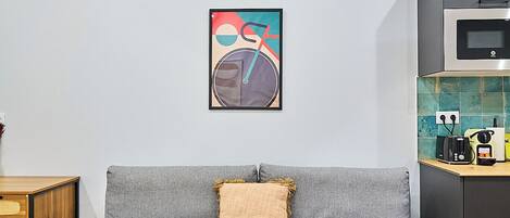 A modern living room with a grey sofa, a wooden floor, a kitchen area with a microwave, and a colorful abstract painting on the wall.