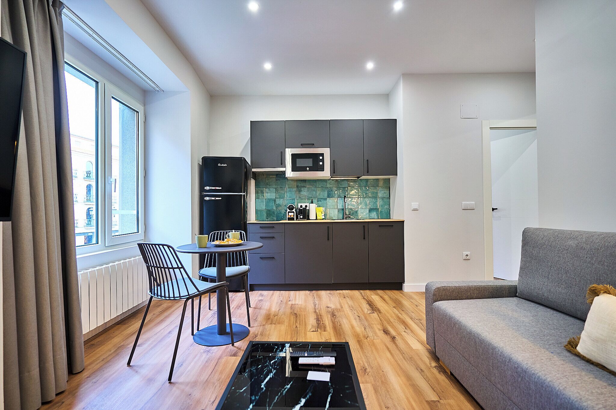 A modern living room with a grey sofa, a wooden floor, a dining table with chairs, and a kitchen area with dark cabinets and a microwave.