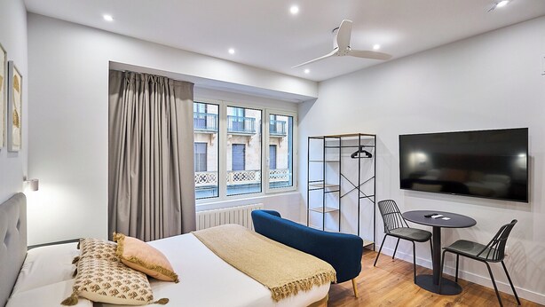 Een moderne hotelkamer met een bed, een televisie, een eettafel met stoelen en een raam met uitzicht op gebouwen.