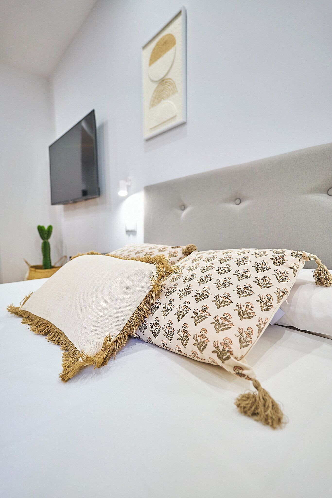A neatly made bed with patterned pillows and a decorative throw, a flat-screen TV mounted on the wall, and a small potted plant on the nightstand.