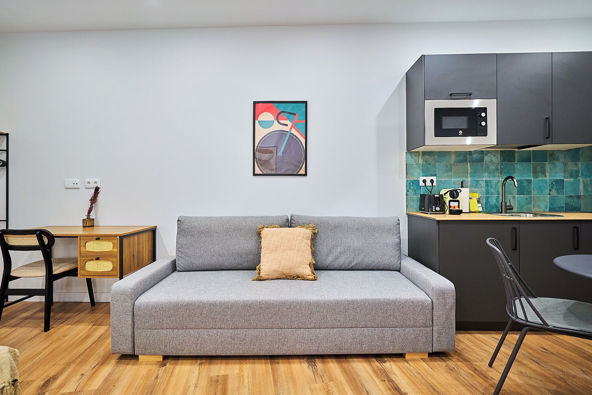Un espacio habitable compacto con un sofá gris, un escritorio de madera, una zona de comedor y una cocina equipada con microondas.