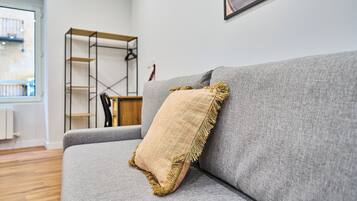 A grey sofa with a beige cushion and a wooden shelf unit against a white wall.