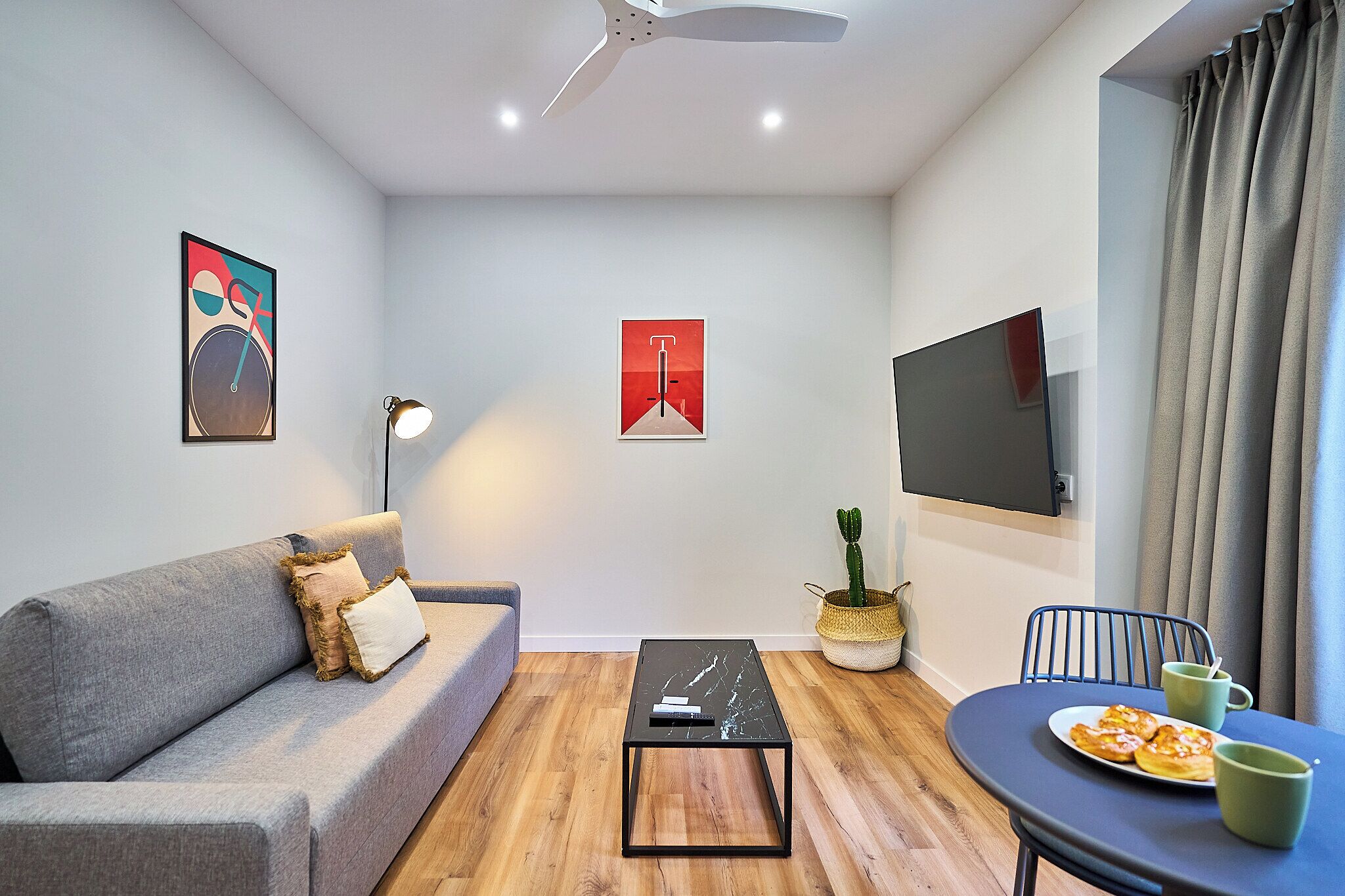 Una sala de estar moderna con un sofá gris, una mesa de centro negra, una mesa de comedor con un plato de comida y tazas verdes, una planta de cactus y un televisor de pantalla plana montado en la pared.