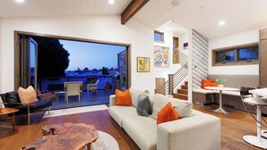 A spacious living area with a large window, a sofa, a coffee table, and a dining area with a chair and table. - Ocean View Craftsman Home - Great Location + Walk to Beach (Encinitas)