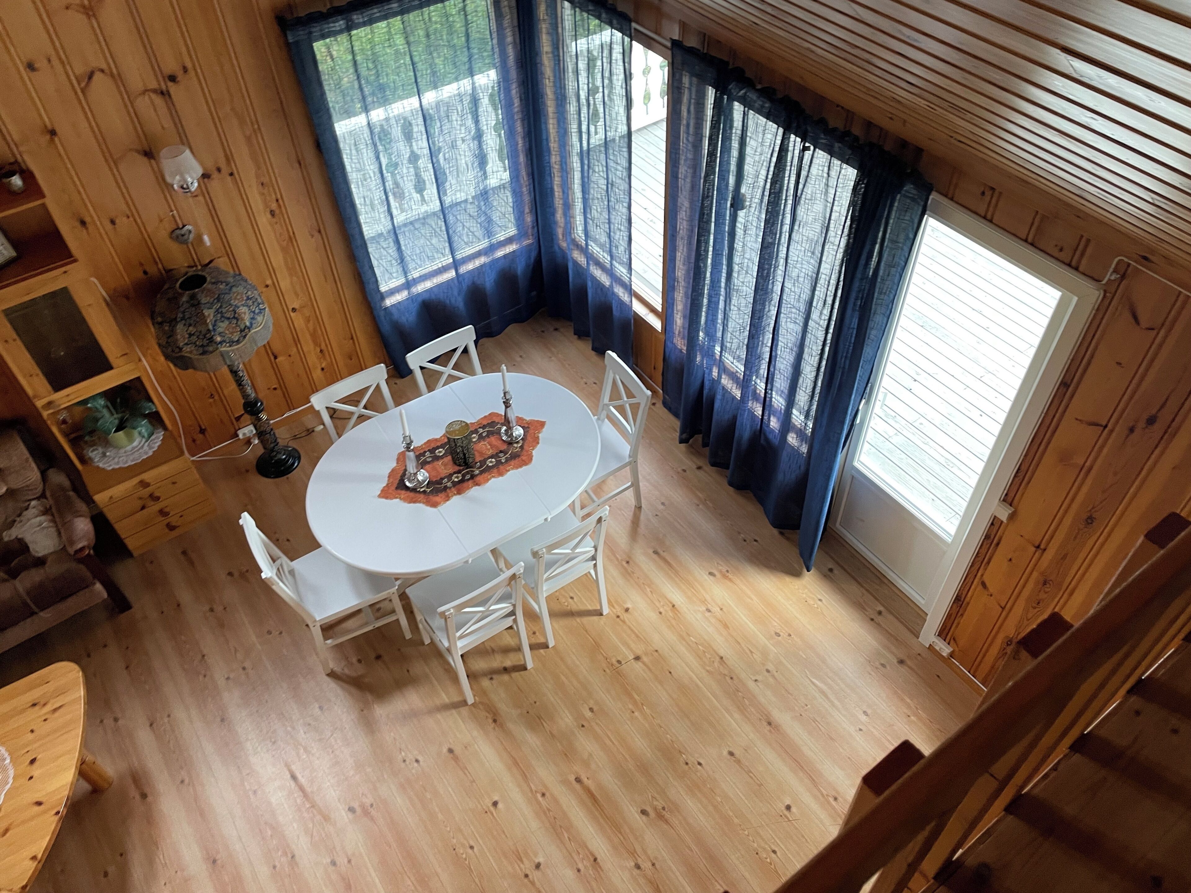 A wooden-paneled room with a round table and white chairs, a small shelf with decorative items, and a window with blue curtains.