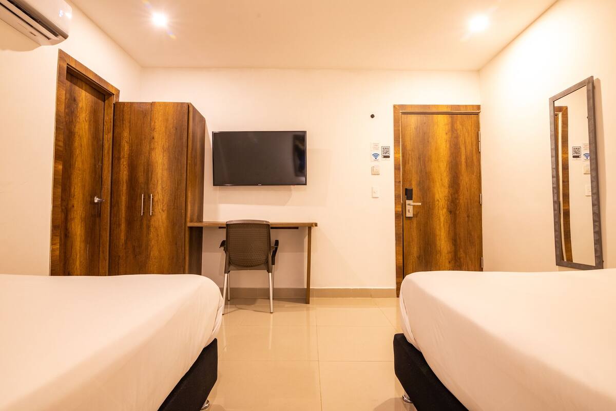 A hotel room with two beds, a desk, a chair, a TV, and wooden wardrobes.