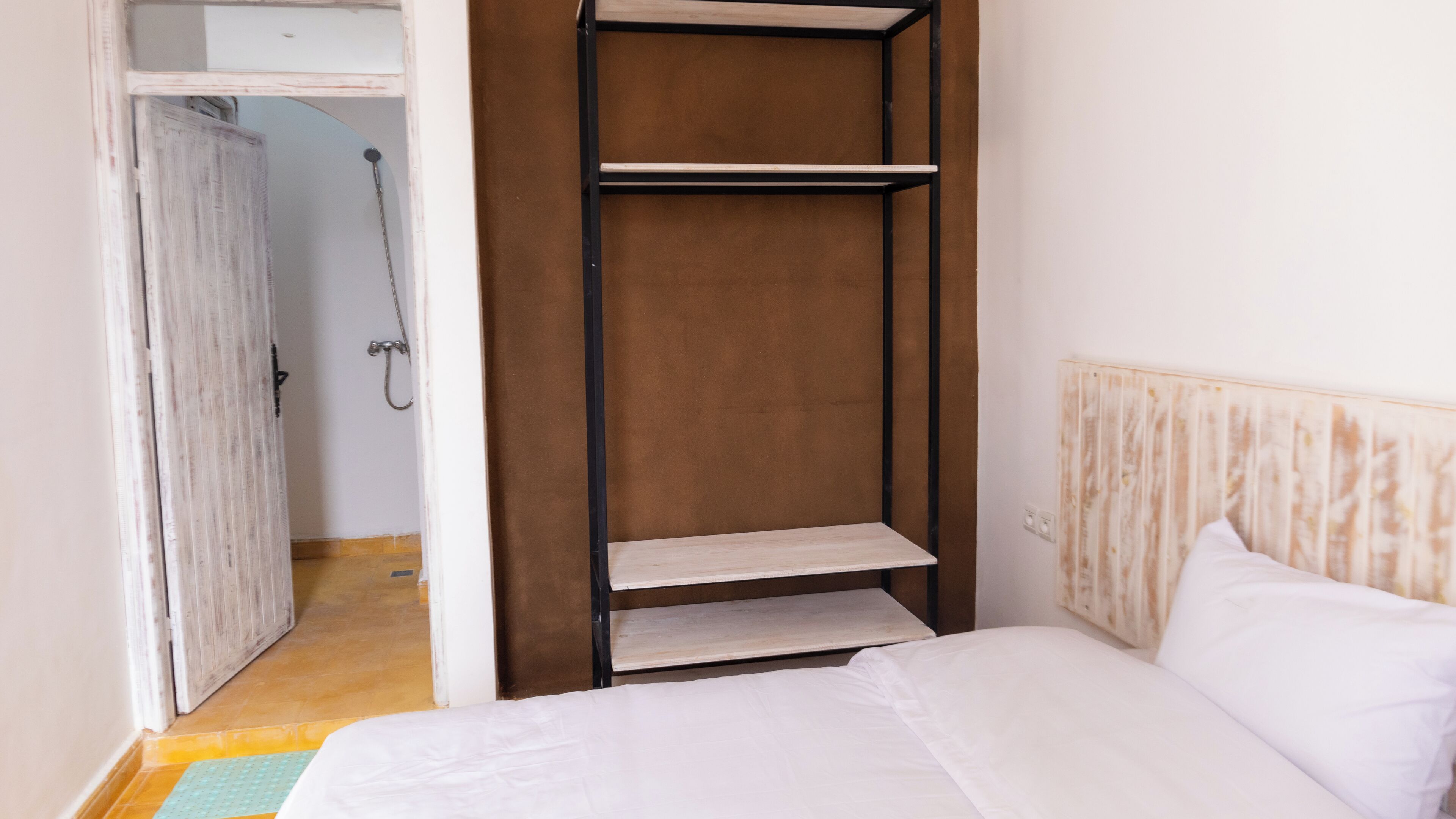 Un dormitorio con cama, cabecera de madera, armario con estantes y baño visible a través de una puerta abierta.