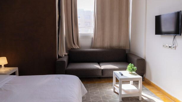 Une chambre d’hôtel avec un lit, un canapé, une petite table avec une plante, un téléviseur fixé au mur et une fenêtre avec des rideaux.