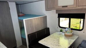 A compact RV interior with a kitchenette, a dining area, a small seating space, and a window providing a view of the outdoors. - Cozy Camper in Charming Whittier with Heat & AC (Whittier)