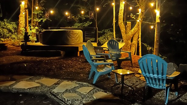 An outdoor seating area with chairs and tables, surrounded by trees and illuminated by string lights. - Cozy Camper in Charming Whittier with Heat & AC (Whittier)