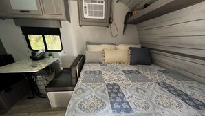 A compact RV interior with a bed, a small table, and a window. - Cozy Camper in Charming Whittier with Heat & AC (Whittier)