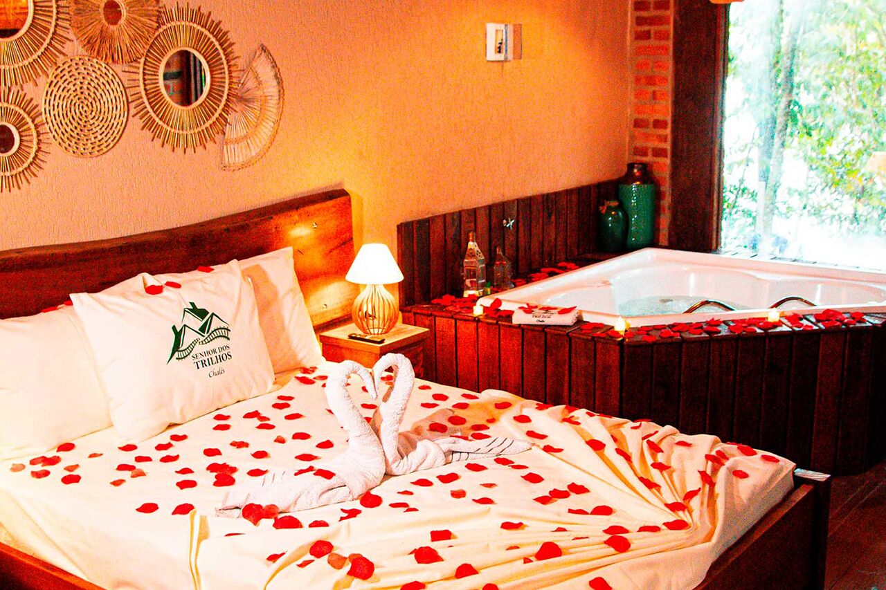 A hotel room with a bed adorned with rose petals and a swan-shaped towel, a wooden headboard, a wall-mounted mirror, and a window with a view of greenery.