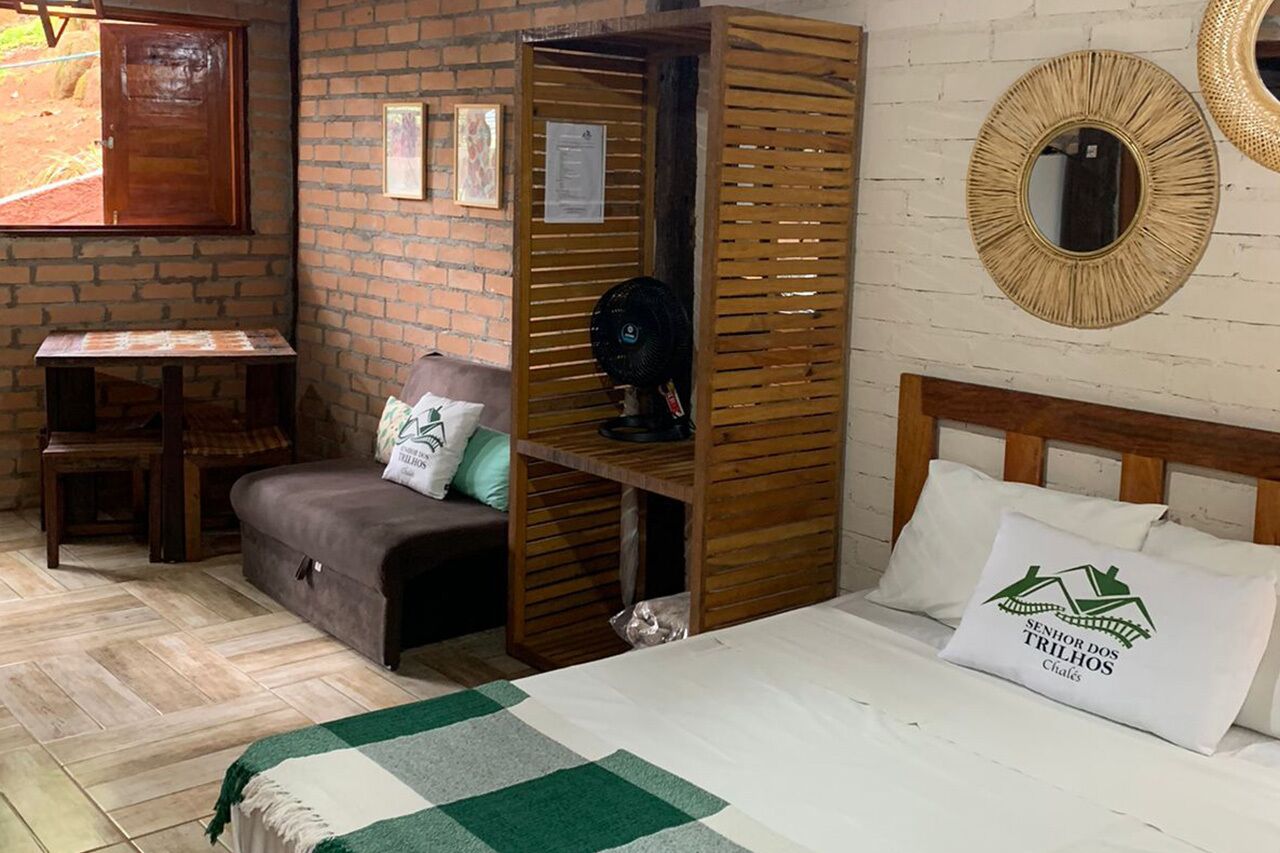A room with a bed, a wooden bench, a small table, and a brick wall.