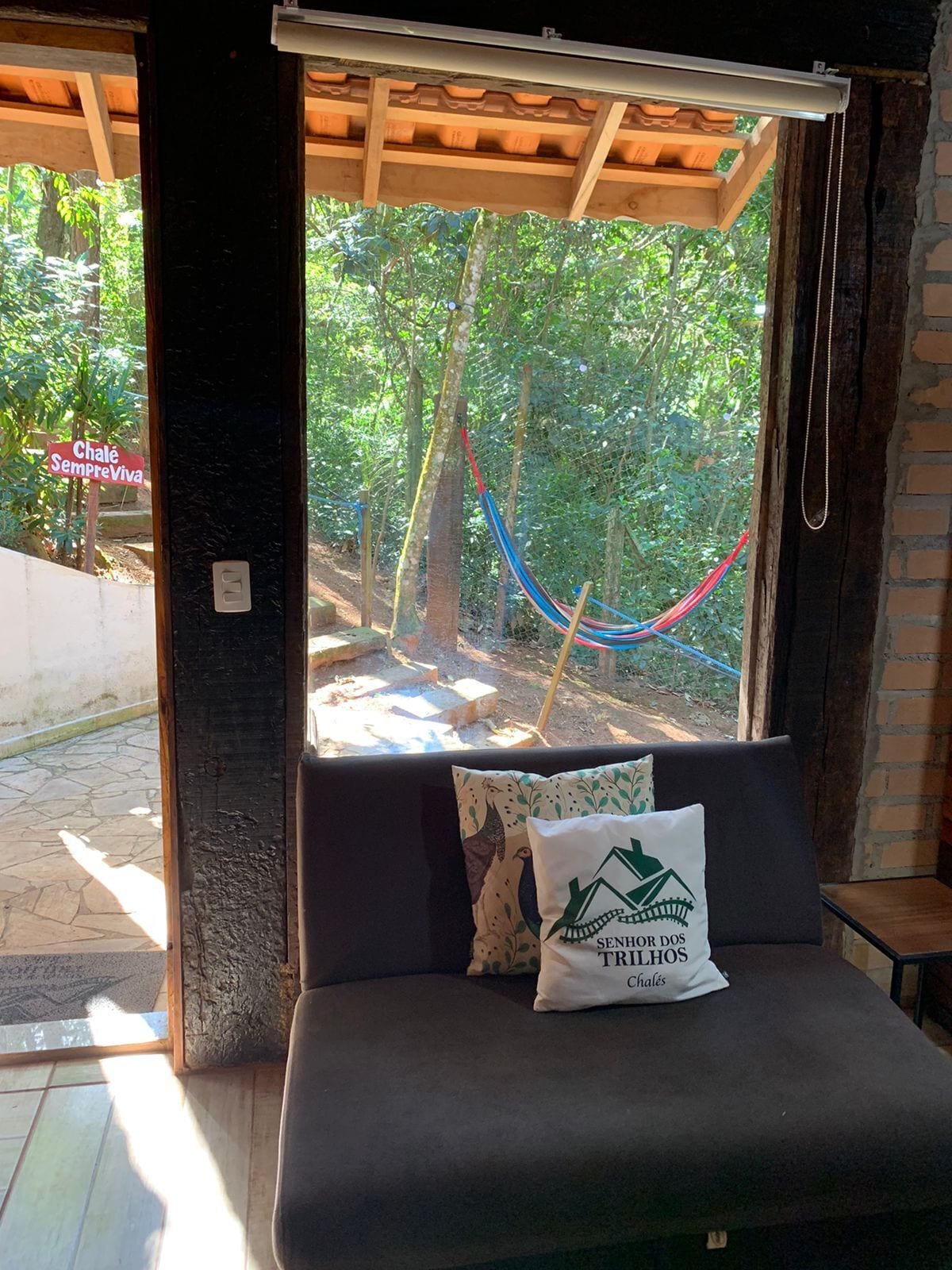 Quarto com vista para uma rede entre árvores, almofada com logotipo e um banco de madeira.