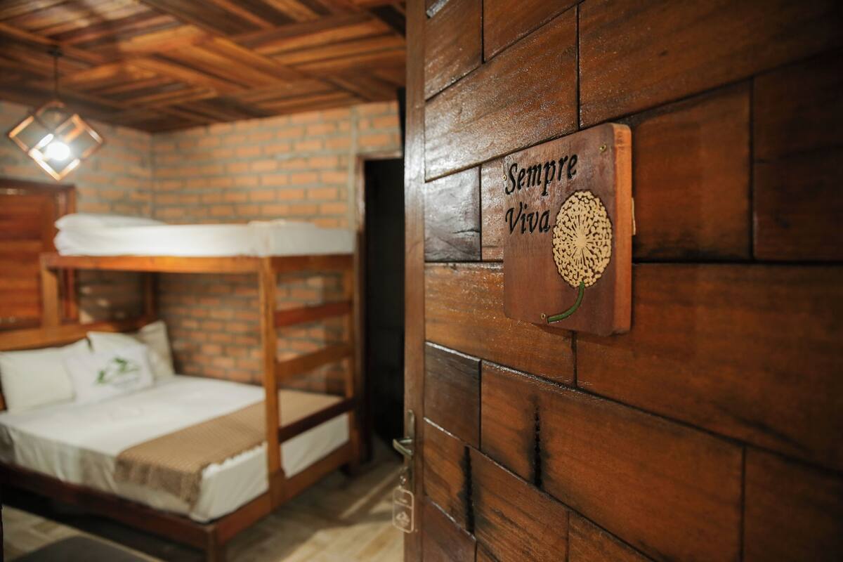 A wooden bunk bed with a white mattress and pillows, a wooden wall with a sign that reads "Sempre Viva," and a hanging light fixture.