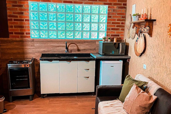 A compact kitchen with a brick wall, a window with a grid pattern, a microwave, and a small refrigerator. - Chales senhor dos trilhos (Mairipora)