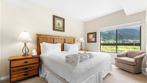 Une chambre à coucher avec un grand lit, deux tables de chevet, une chaise et une fenêtre donnant sur un paysage verdoyant.