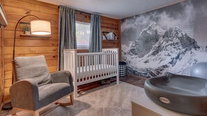 Et hyggeligt værelse med en vugge, en gyngestol, en vægmonteret hylde med bøger, et stort maleri af et bjerglandskab og et babybadekar.