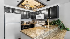 A modern kitchen with dark cabinets, granite countertops, a white refrigerator, a white microwave, a stainless steel sink, and a ceiling light fixture.