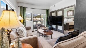 A modern living room with a sofa, armchair, and coffee table, a TV, and a balcony with a view of mountains.