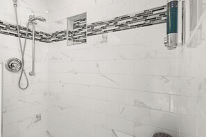 Un baño con baldosas de mármol blanco, una ducha con cabezal de ducha manual y una barra vertical con una botella.