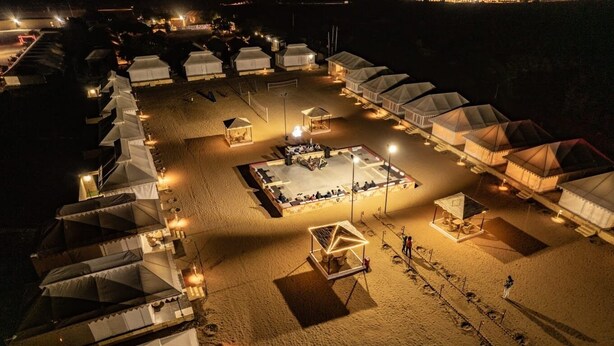 An aerial view of a beachside resort with multiple tents arranged in rows, a central open area with a seating arrangement, and a sandy beach.