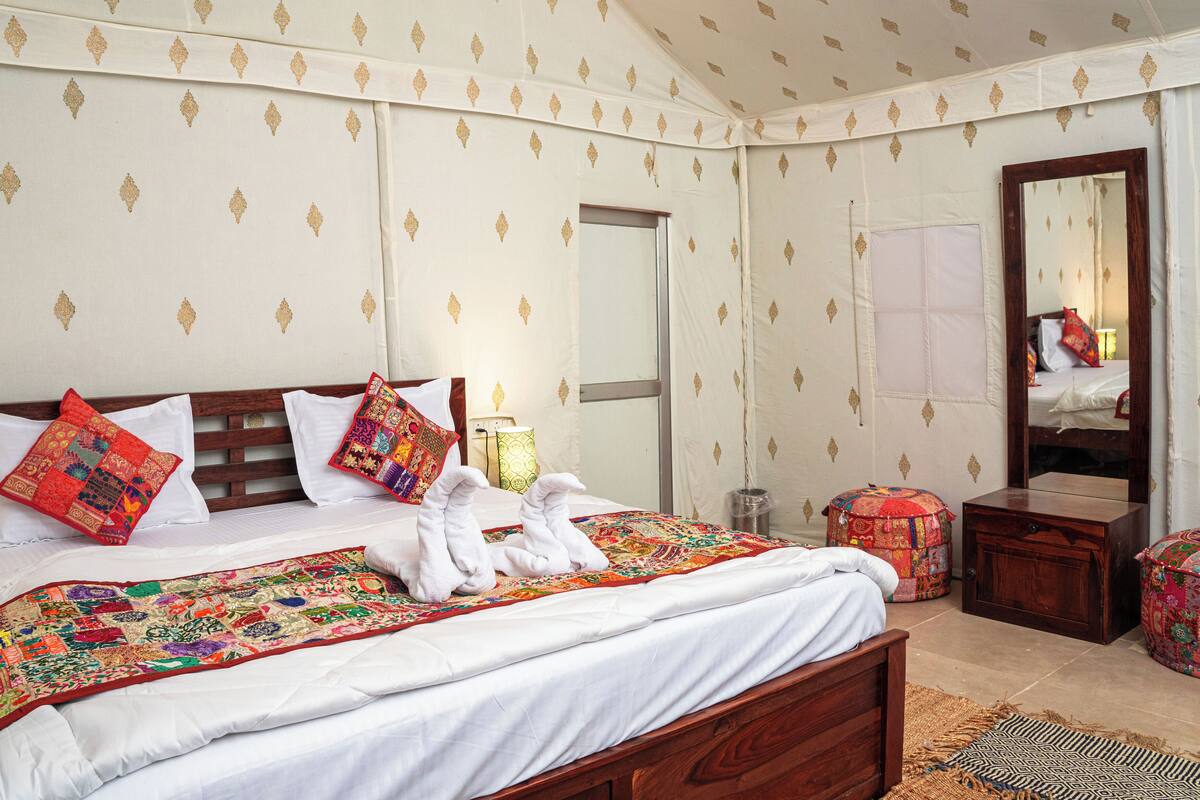 A neatly made bed with a colorful quilt, a wooden headboard, and decorative pillows. There is a small bedside table with a lamp and a mirror on the wall.