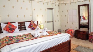 A neatly made bed with a colorful quilt, a wooden headboard, and decorative pillows. There is a small bedside table with a lamp and a mirror on the wall.