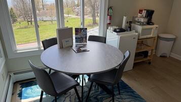 Una mesa redonda con cuatro sillas, un refrigerador pequeño, un microondas y un basurero en una habitación con un ventanal grande.