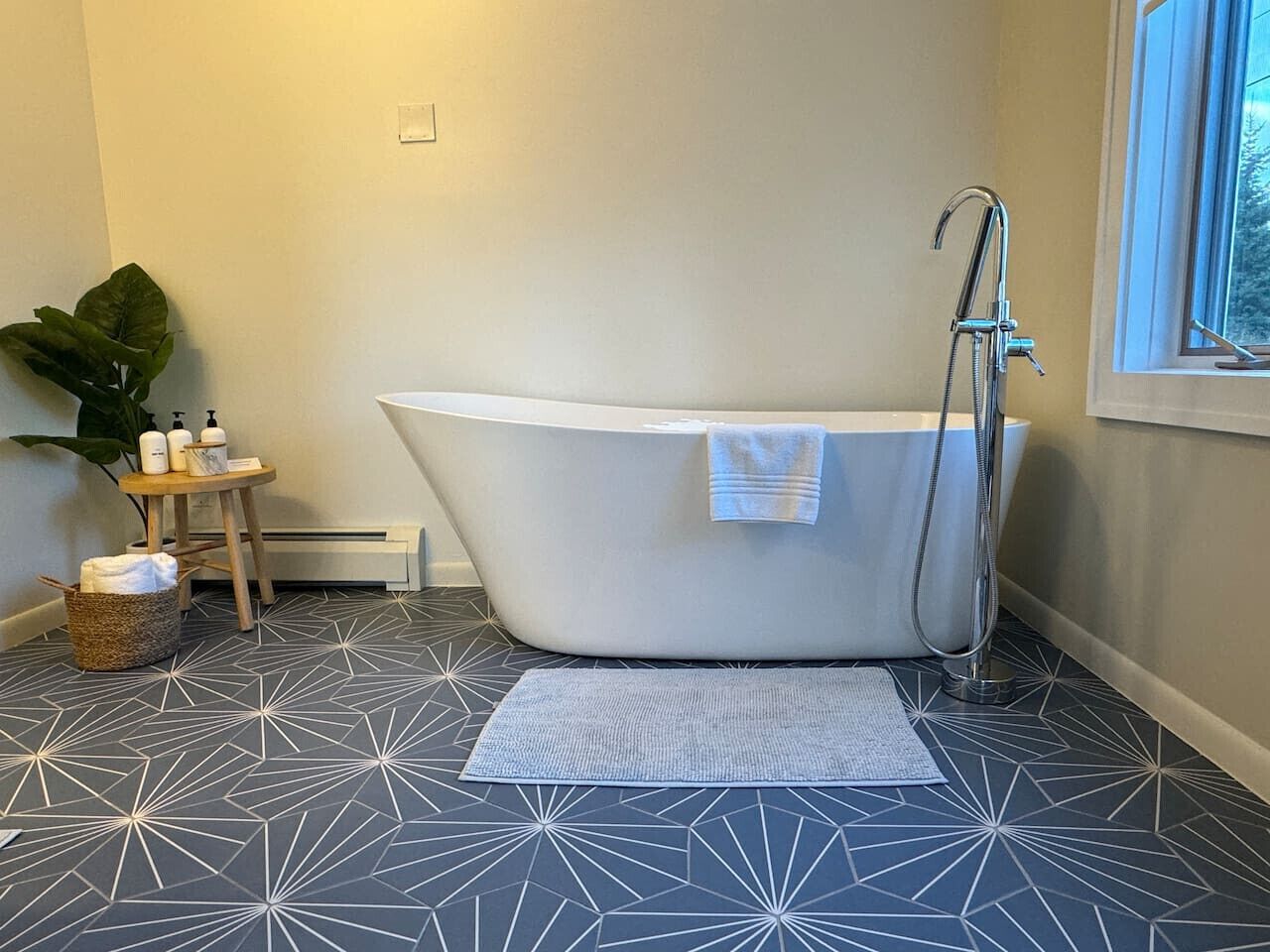 Un bagno moderno con una vasca freestanding, un piccolo sgabello di legno, una pianta e un pavimento decorato.