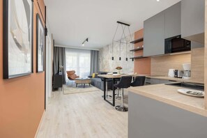 Et moderne køkken med en central ø, et spiseområde med sofa og stole, og et stort vindue der giver naturligt lys.